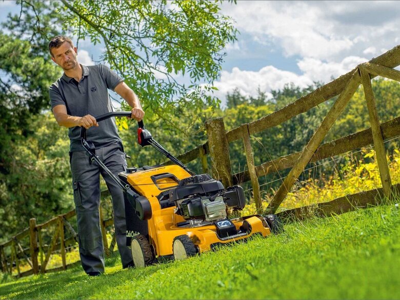 Person mäht mit einem Cub Cadet Benzin-Rasenmäher auf einem grünen Grundstück mit Holzzaun.